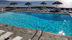 A pool with clear blue water, surrounded by white lounge chairs and thatched umbrellas, with a view of the sea and distant hills. - COZY 2 BEDROOM OCEAN VIEW las gaviotas  17#2 (rosarito beach)