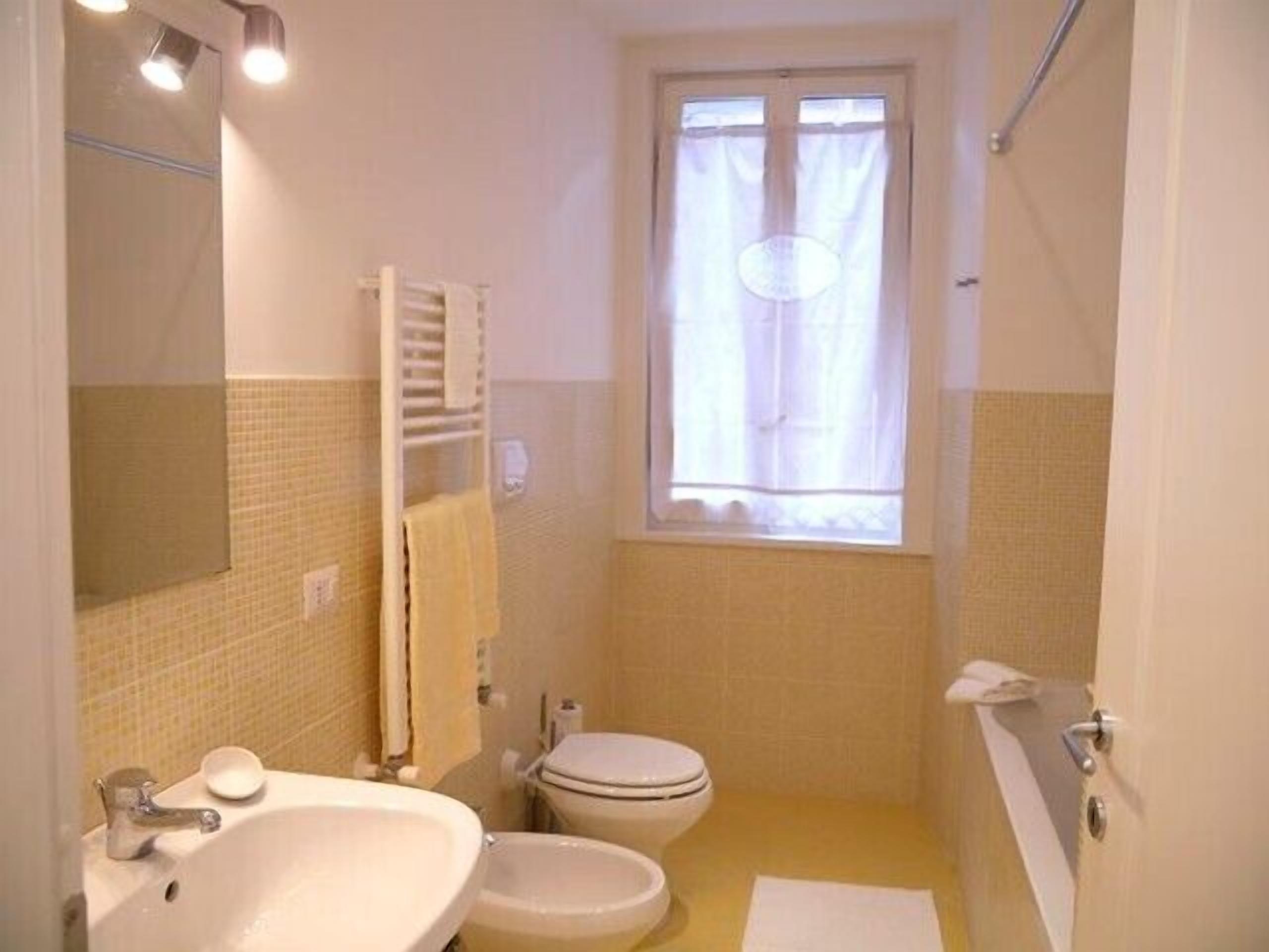 Une salle de bain équipée d’un lavabo, de toilettes et d’un bidet. On y trouve une fenêtre avec des rideaux, un porte-serviettes et un miroir.