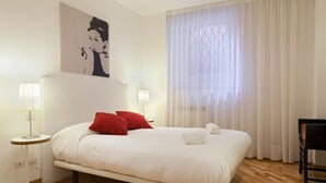 A neatly arranged bedroom with a bed, two red pillows, a white blanket, a bedside table with a lamp, and a chair. - Fontana di Trevi and Spanish Steps. Modern and elegant (Roma)