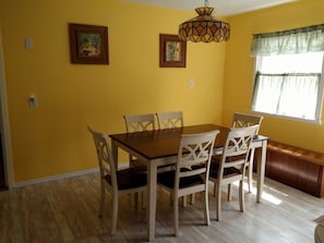 Un comedor con una mesa de madera y sillas blancas, paredes amarillas y dos cuadros enmarcados.