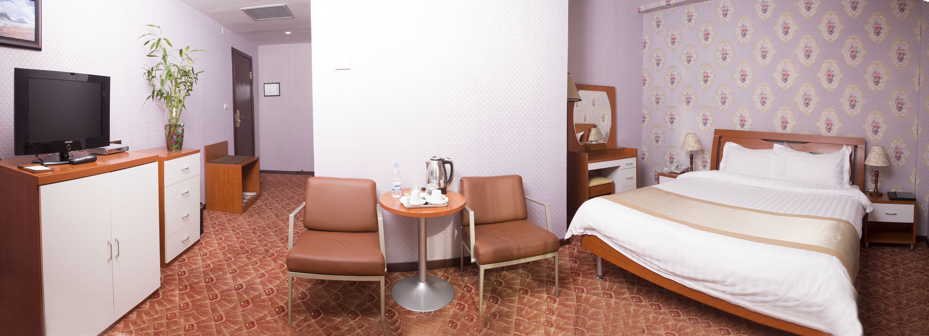 A hotel room with a bed, a desk with a computer, a chair, a small table with a kettle, and a TV.