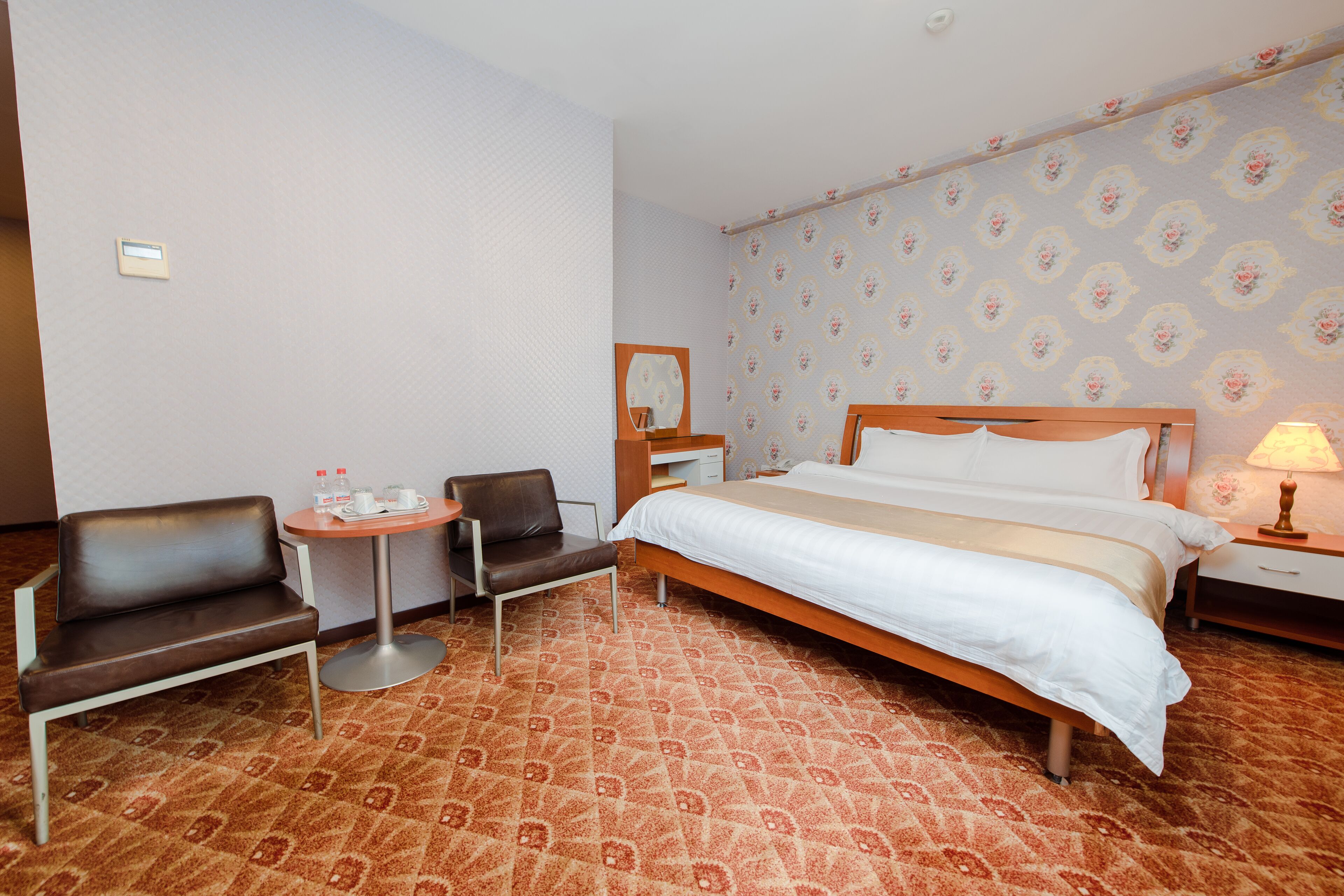 A hotel room with a bed, two chairs, a small table, a nightstand, and a lamp.