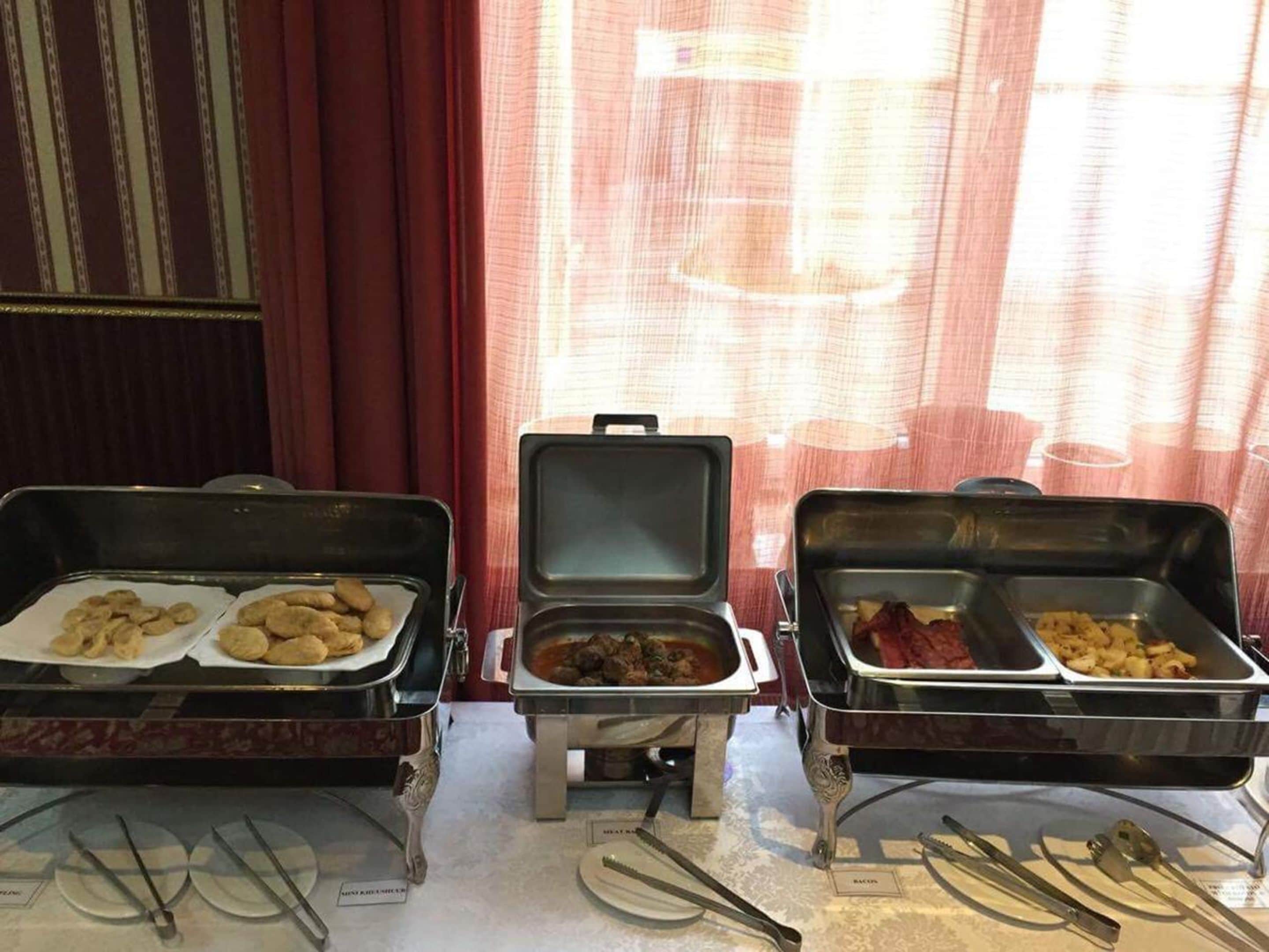 Servicio de comidas tipo buffet con diversos platos en recipientes metálicos, dispuestos sobre una mesa con cubiertos y platos.