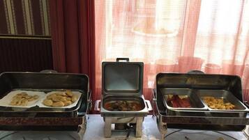 Servicio de comidas tipo buffet con diversos platos en recipientes metálicos, dispuestos sobre una mesa con cubiertos y platos.