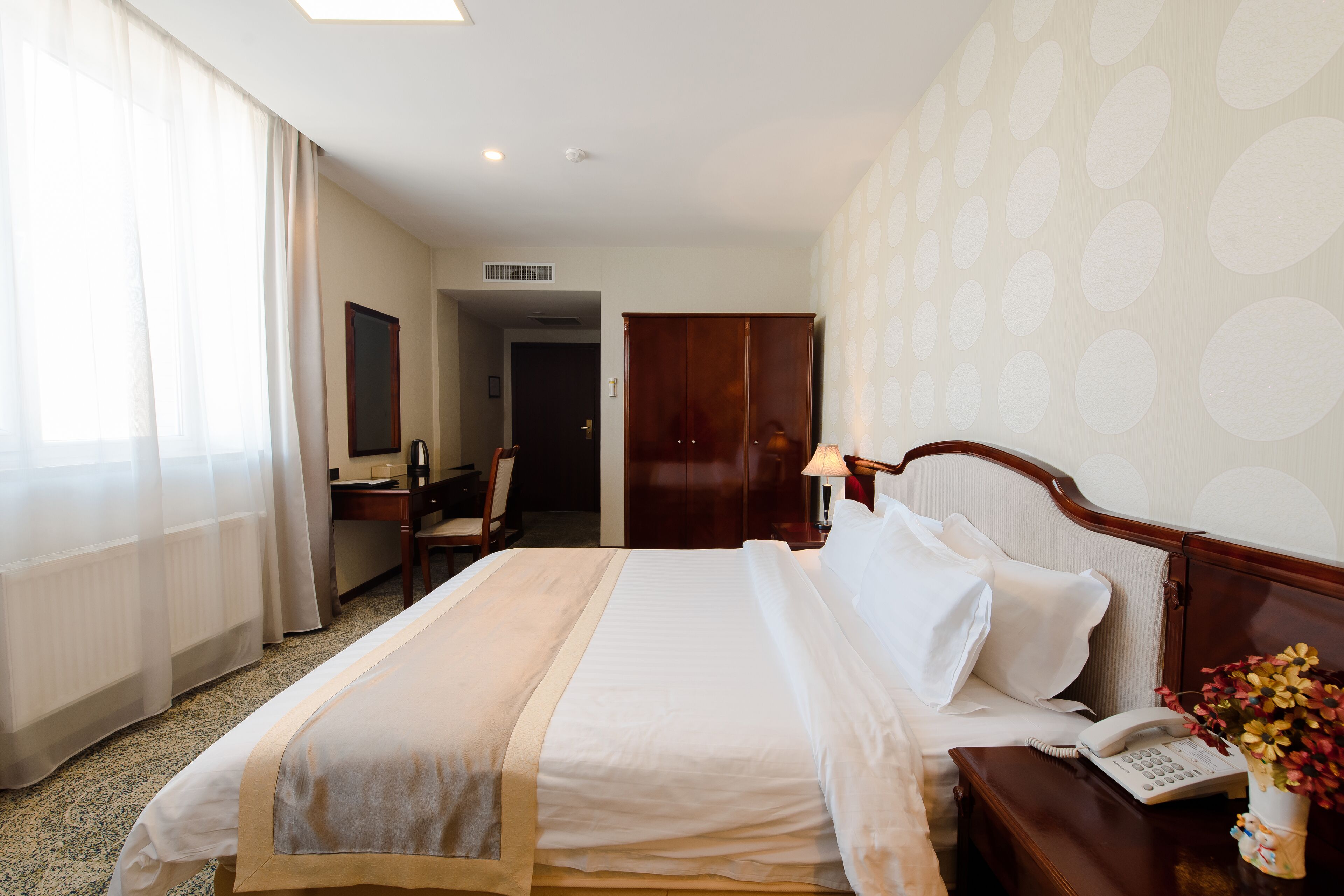 Une chambre d’hôtel avec un lit bien fait, un bureau avec une chaise, une armoire et une table de chevet avec une lampe et un téléphone.