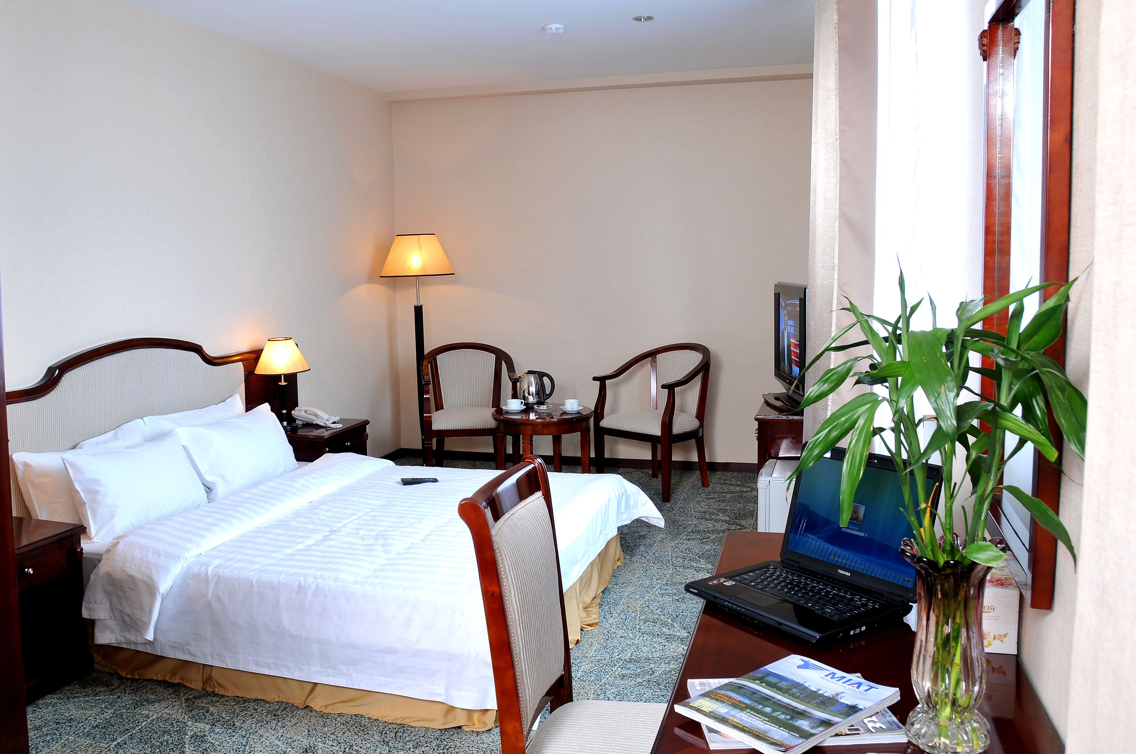 Habitación de hotel con cama, escritorio, laptop, silla, lámpara y planta en maceta.