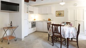 Una cocina con armarios blancos, una mesa de madera con sillas, un televisor montado en la pared y un reloj en la pared.