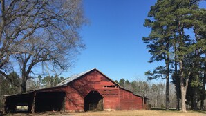 Ein rotes Bauernhaus mit einer großen offenen Tür und einem überdachten Ladebereich, umgeben von Bäumen und blauem Himmel.