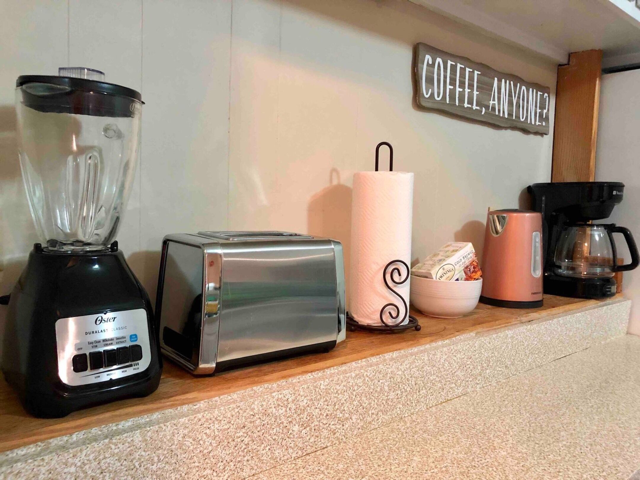 A kitchen counter with a blender, toaster, coffee maker, and a sign that reads "COFFEE, ANYONE?".