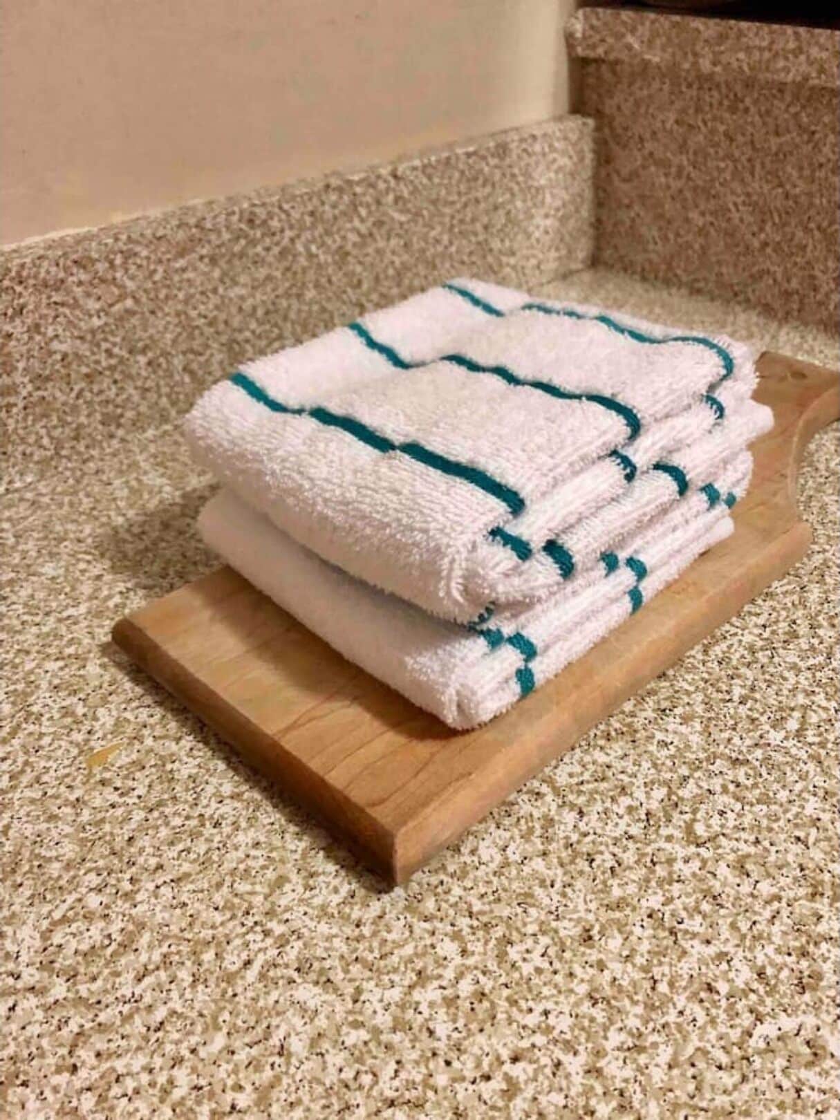 En hög med vita handdukar med turkos kant, placerad på ett träbricka som står på en mönstrad matta.