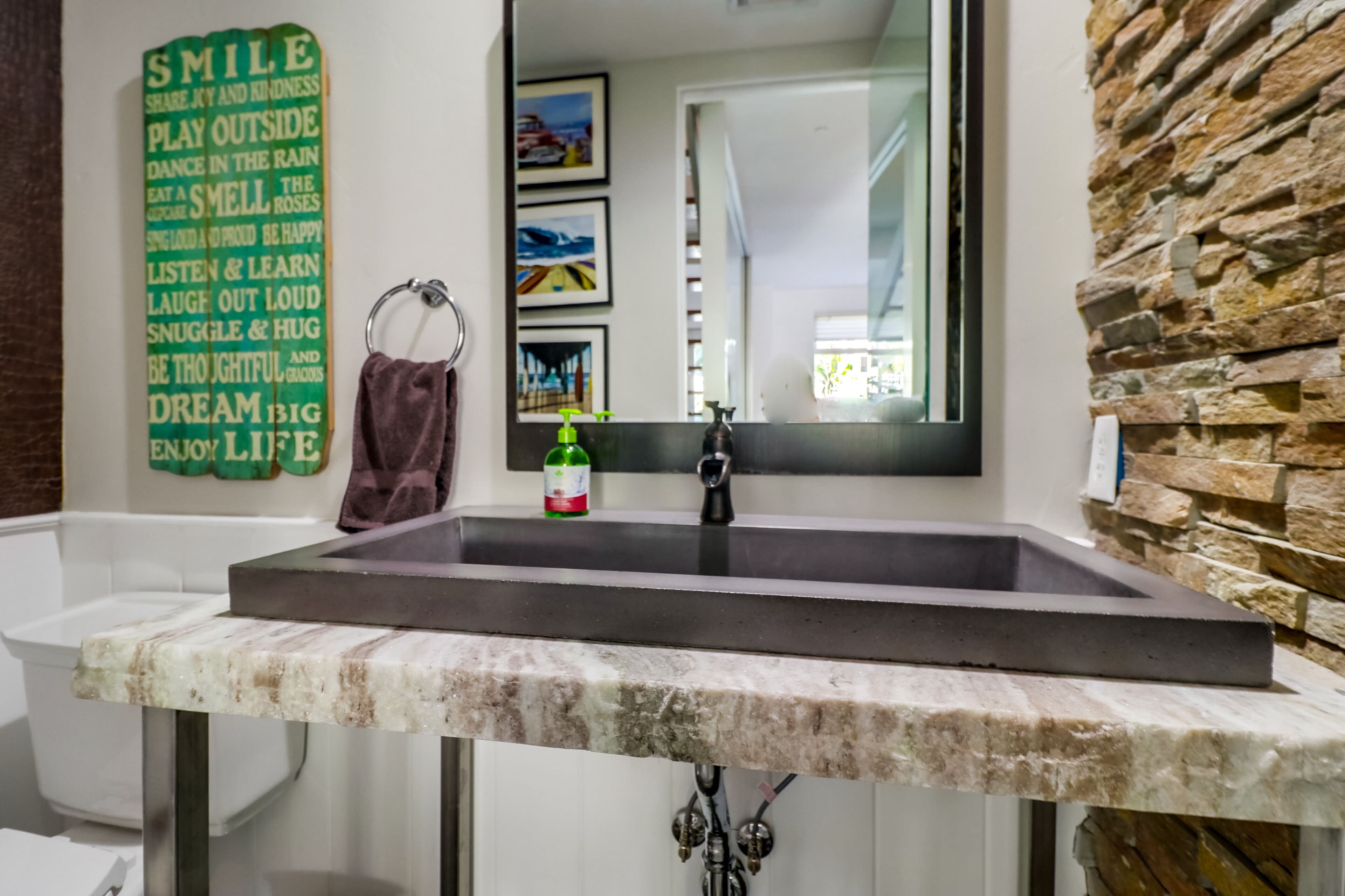 A bathroom with a marble sink, a stone wall, a framed motivational quote, and a mirror reflecting a room with a window.