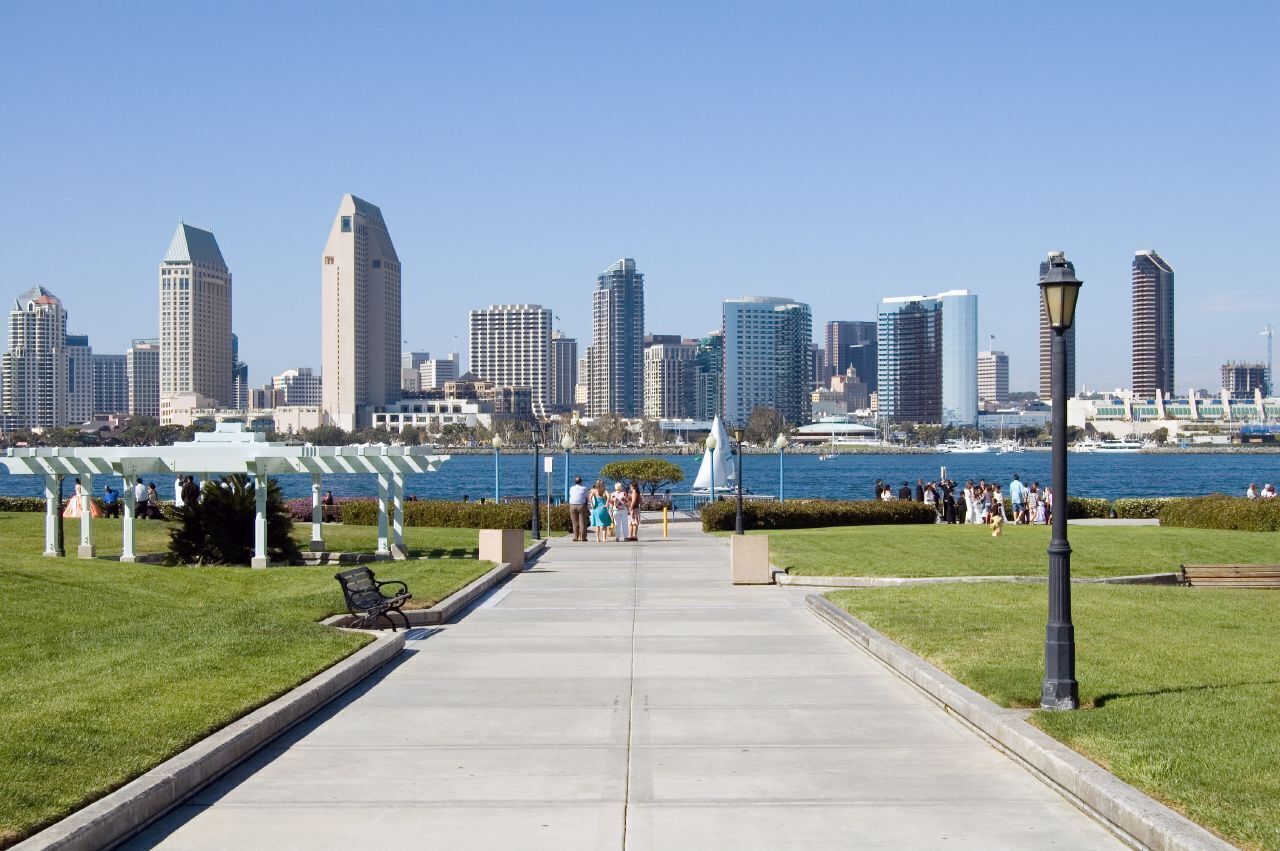 Un paseo marítimo con el horizonte de la ciudad al fondo, bancos y un cielo despejado.