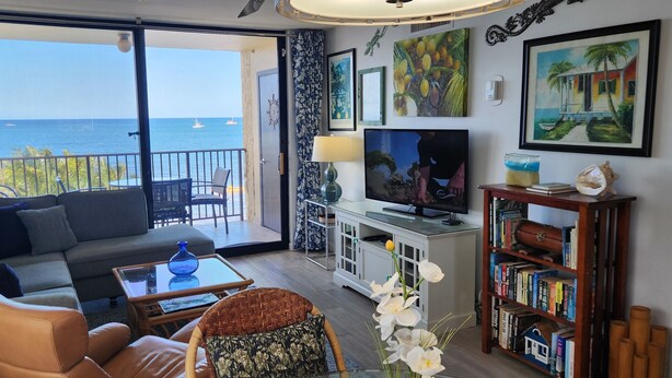 Un salon comprenant un canapé, des fauteuils, une table basse, un téléviseur, une bibliothèque et un balcon avec vue sur la mer.