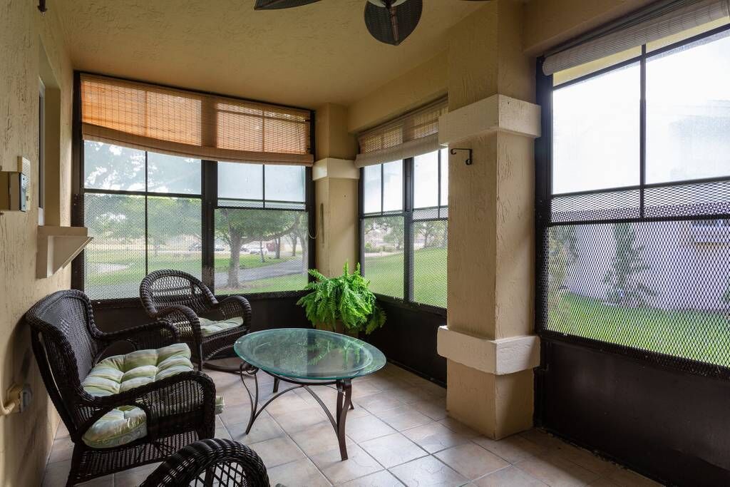 Ein überdachter Patio-Bereich mit Korbmöbeln und einem Glastisch, große Fenster mit Jalousien und Blick auf Grünfläche.
