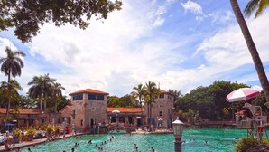 Une grande piscine extérieure entourée de palmiers et de bâtiments aux toits recouverts de tuiles rouges.