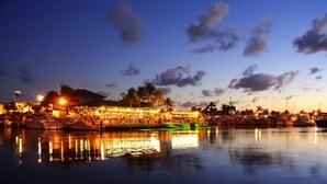 Un hôtel situé au bord de l’eau, doté d’une marina, où les lumières se reflètent sur l’eau, avec des palmiers et un ciel au coucher du soleil.