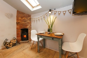 A dining area with a brick fireplace, a table set for two, and a decorative string of heart-shaped flags. - Bothy In Victorian Walled Garden (Worcester)