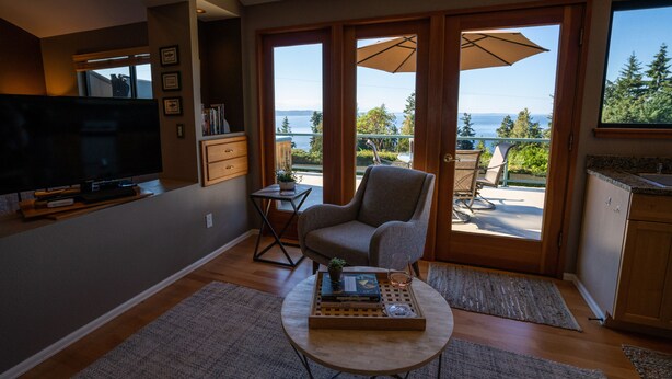 A living room with a TV, a chair, a coffee table with books and a glass, and a view of the outdoors. - Private Cottage and Hot Tub, Spectacular Sunset & Mtn Views (Burien)