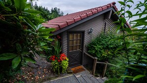 A small, dark wooden house with a red-tiled roof, surrounded by lush greenery and a gravel path leading to the door. - Private Cottage and Hot Tub, Spectacular Sunset & Mtn Views (Burien)