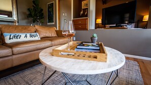 A living room with a brown leather sofa, a coffee table with a tray holding a glass of wine, books, and a small plant. - Private Cottage and Hot Tub, Spectacular Sunset & Mtn Views (Burien)