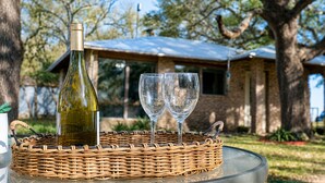 A bottle of wine and two glasses on a wicker tray, with a house and trees in the background. - Peaceful Farm Cabin for Couples | TX Hill Country (Georgetown)