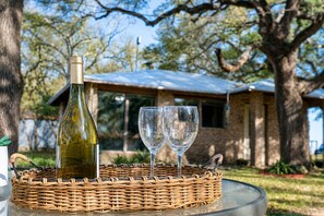 A bottle of wine and two glasses on a wicker tray, with a house and trees in the background. - Peaceful Farm Cabin for Couples | TX Hill Country (Georgetown)