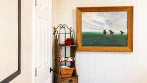 A door with a white frame, a wall-mounted wicker shelf with a red rose, and a framed painting of horses. - Peaceful Farm Cabin for Couples | TX Hill Country (Georgetown)