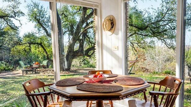 A round wooden table with woven placemats, surrounded by wooden chairs, set against a large window overlooking a garden with trees and a patio area. - Peaceful Farm Cabin for Couples | TX Hill Country (Georgetown)