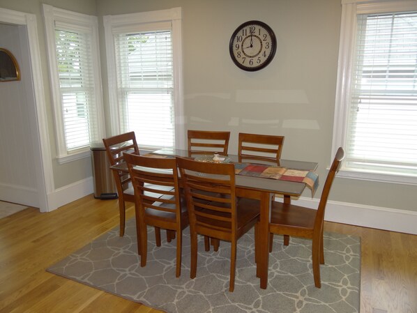 Comedor con mesa y sillas de madera, un reloj en la pared y ventanas con persianas.