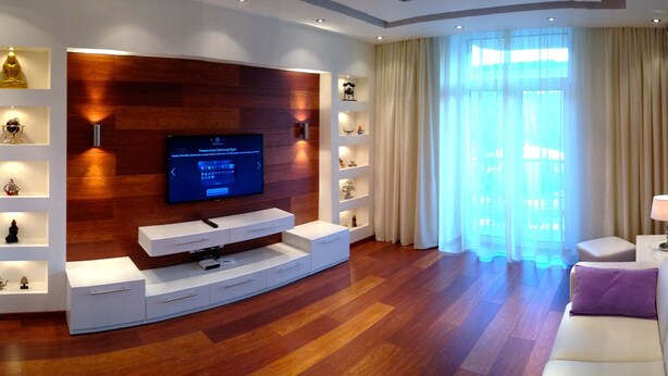 A modern living room with a flat-screen TV, built-in shelves with decorative items, a white sofa, and a large window with sheer curtains.