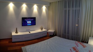 A modern hotel room with a flat-screen TV mounted on the wall, a white bedside table with a statue, a white bed, and sheer curtains.