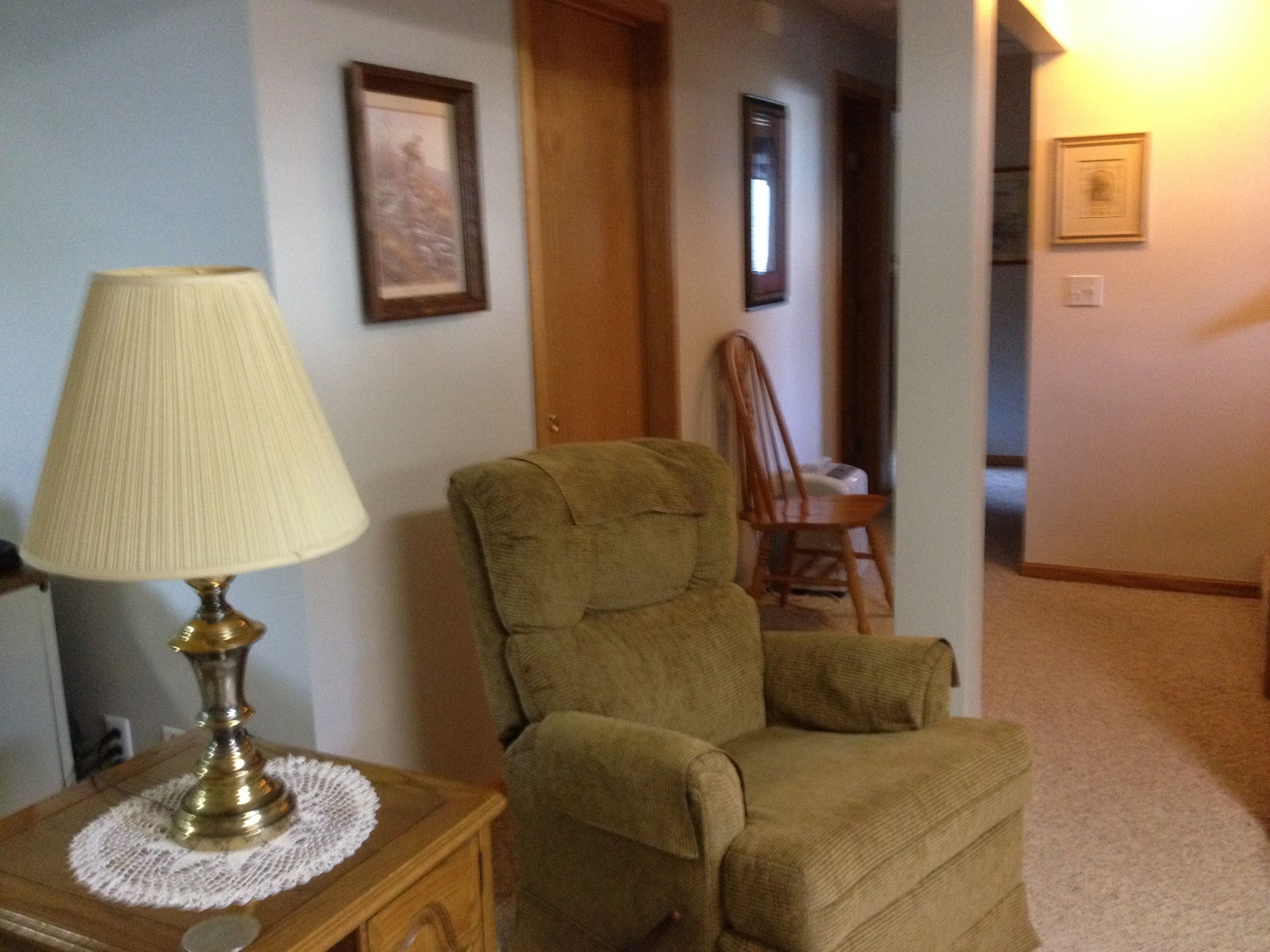 A room with a green armchair, a lamp on a wooden side table, and framed pictures on the walls.