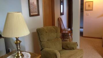 A room with a green armchair, a lamp on a wooden side table, and framed pictures on the walls.