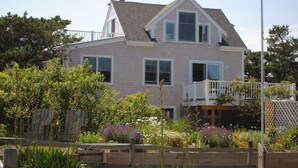A two-story house with a balcony, surrounded by a garden with various plants and flowers. - Ocean Cottage A/C near PB and Beachcomber. Steps to the beach! (Wellfleet)