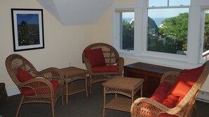 A room with wicker furniture, a framed picture on the wall, and a view of the sea through large windows. - Ocean Cottage A/C near PB and Beachcomber. Steps to the beach! (Wellfleet)