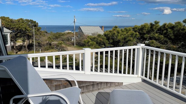 A deck with two lounge chairs, a white railing, and a view of the ocean and trees. - Ocean Cottage A/C near PB and Beachcomber. Steps to the beach! (Wellfleet)