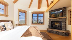Une chambre dotée d’un grand lit, d’une cheminée en pierre et de nombreuses fenêtres offrant une vue sur les montagnes.