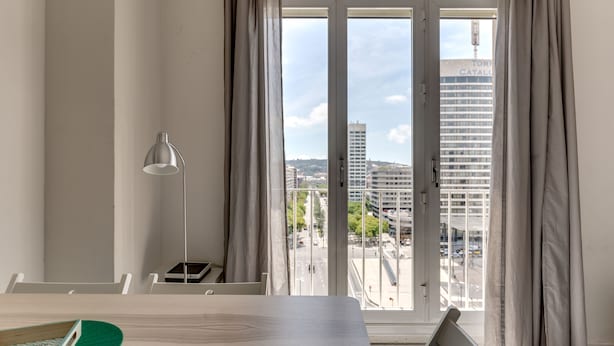 Ein modernes Zimmer mit Esstisch, einer Lampe und Blick auf eine Stadtlandschaft durch ein großes Fenster.