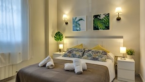 Ein ordentlich gemachtes Bett mit einer braunen Tagesdecke, zwei Kissen und zwei aufgerollten Handtüchern an der Seite. Zwei an der Wand befestigte Nachttische mit Lampen und ein Blumentopf sind ebenfalls vorhanden.