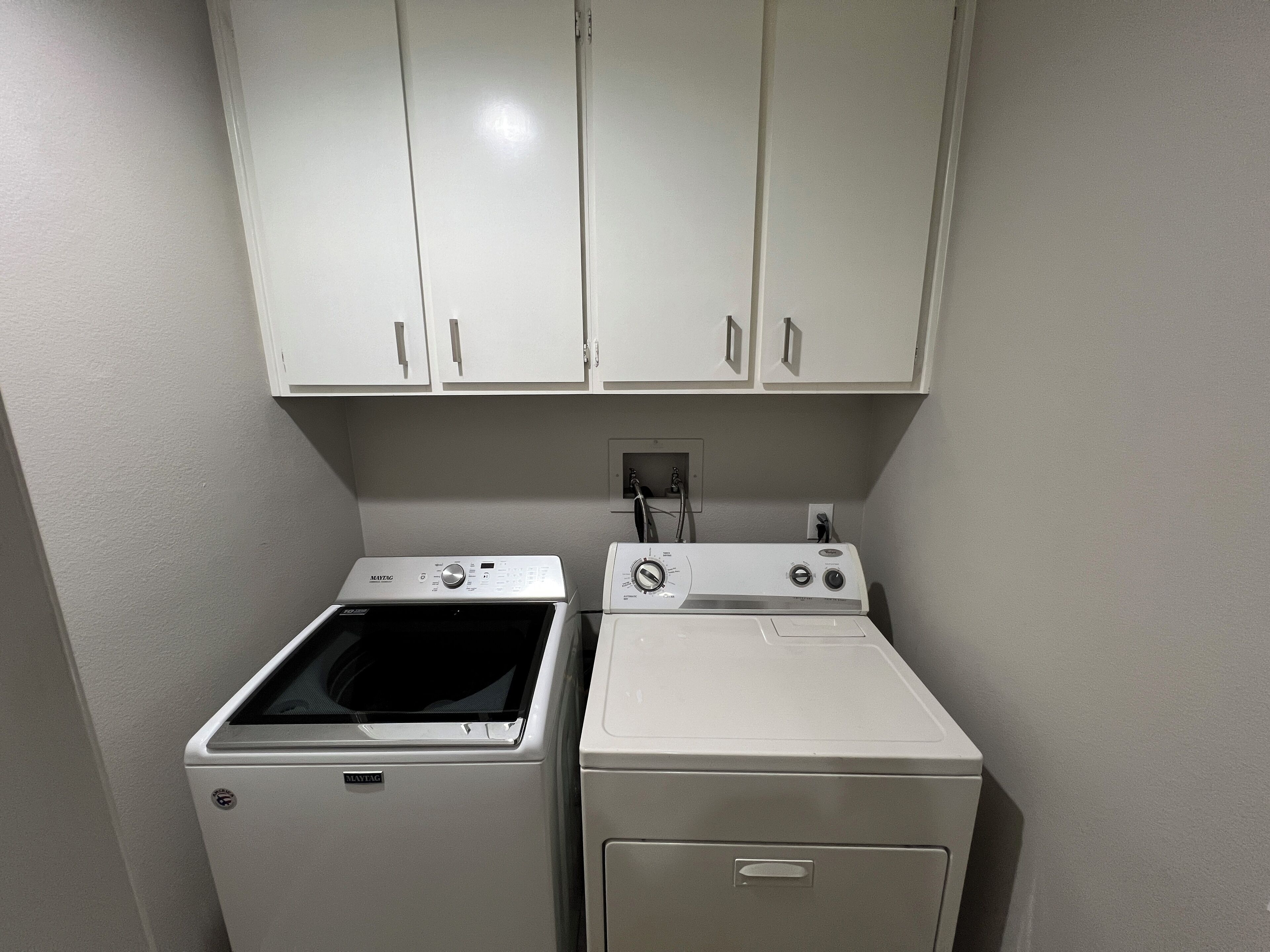 A laundry room with a front-loading washer and dryer, white cabinets above, and a power outlet on the wall.