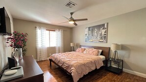 Un dormitorio con cama, lámparas de noche, ventilador de techo, ventana con cortinas y un cuadro en la pared.