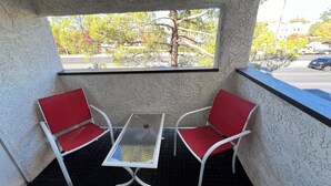 Un balcón con dos sillas rojas y una mesa de vidrio, con vista a una calle con árboles y edificios.