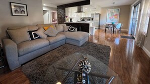 Una sala de estar moderna con un sofá seccional, una mesa de centro de vidrio y una cocina al fondo.