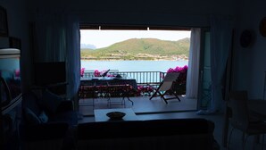 A living room with a view of a lake and hills. There is a TV, a sofa, and a table with chairs.