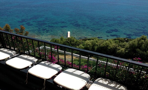 A balcony with white cushioned chairs, a metal railing, and a view of the sea.