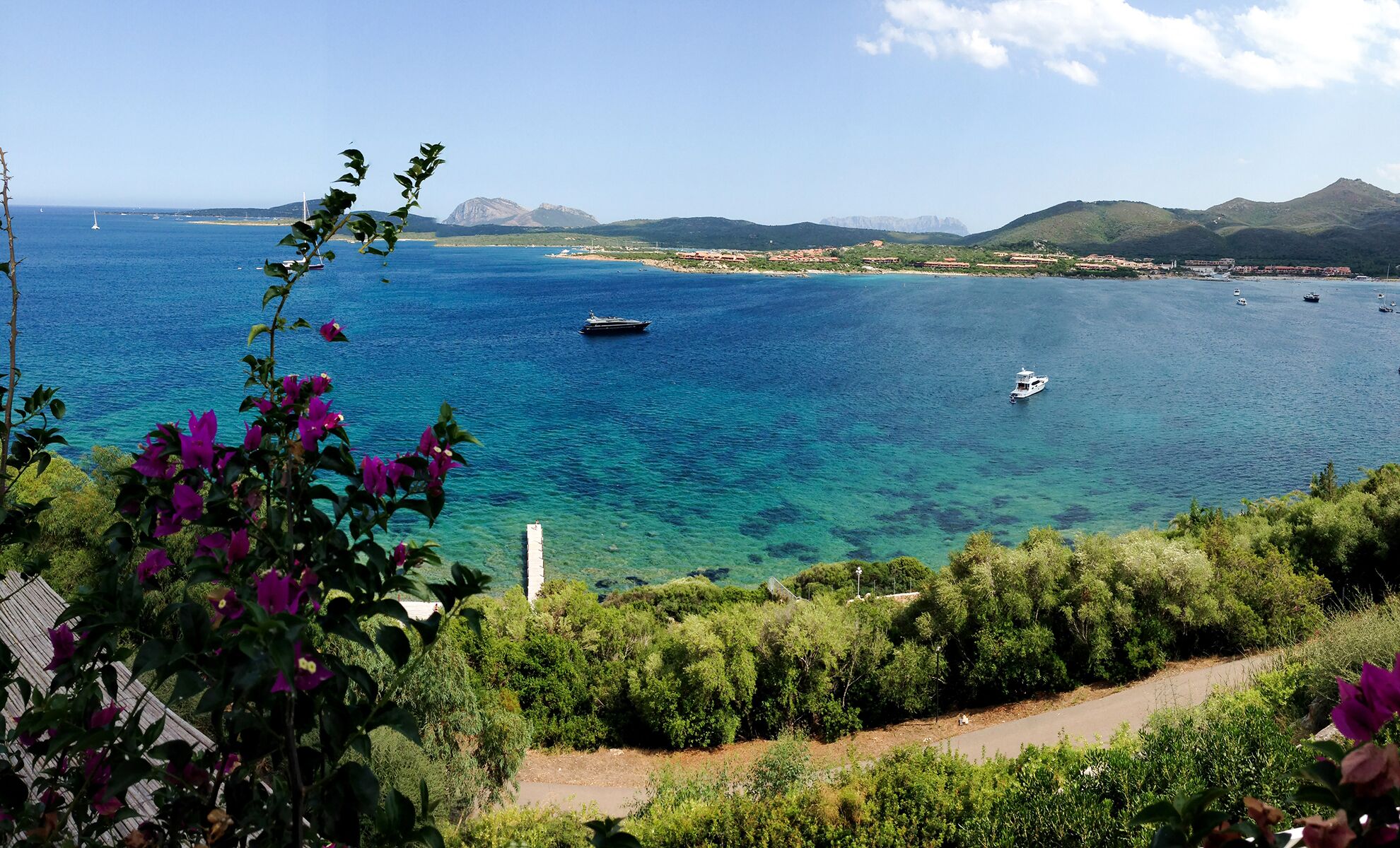 Vista costera con mar azul cristalino, barcos y un paisaje montañoso al fondo.