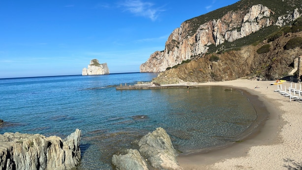 A coastal beach with clear waters, rocky cliffs, and a small island in the distance.