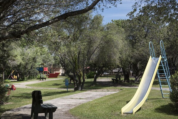 Une aire de jeux avec un toboggan jaune et une échelle bleue, entourée d’arbres et comprenant un espace pique-nique.