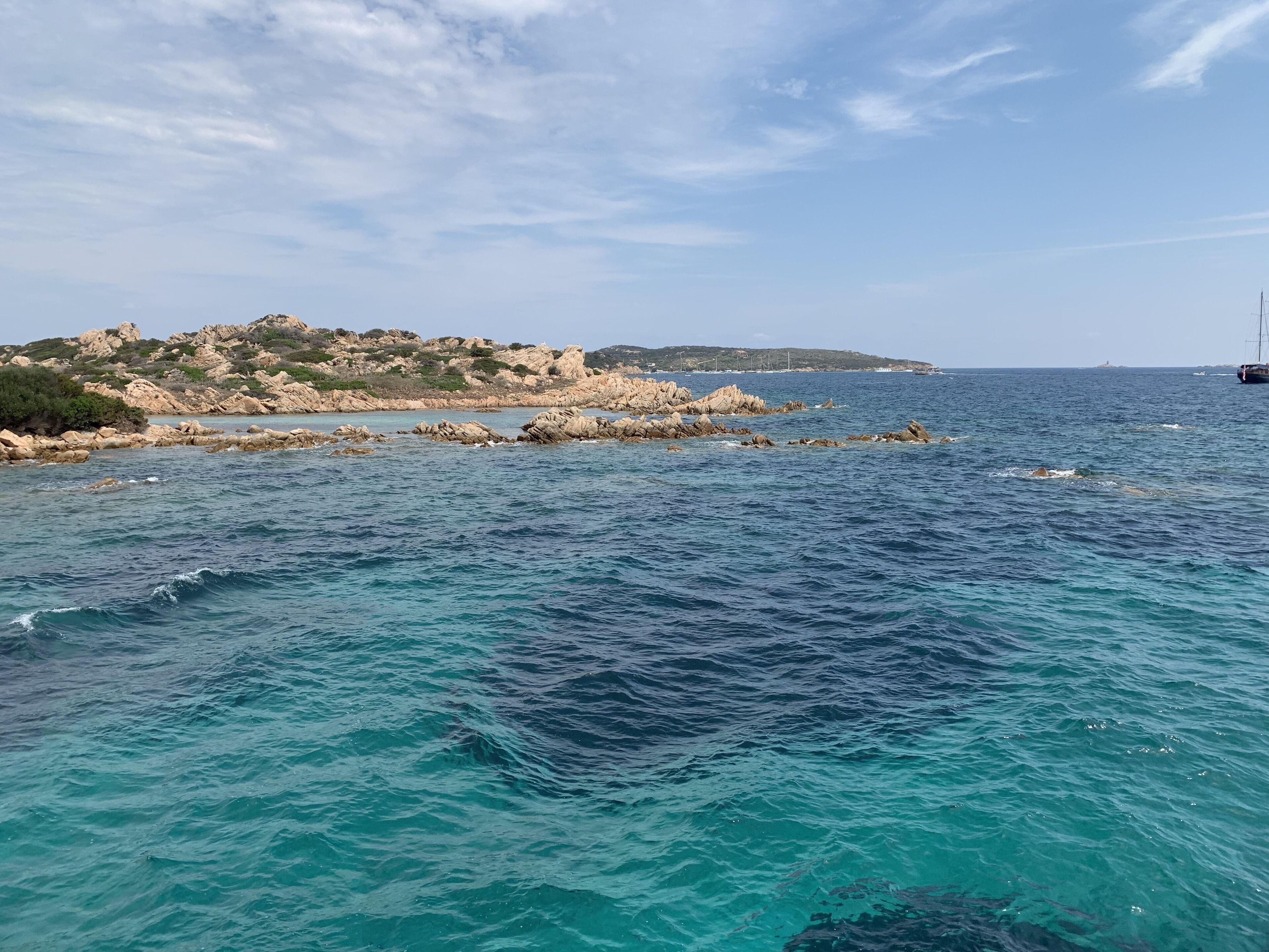 A coastal landscape with clear blue waters, rocky outcrops, and a distant view of a sailboat.