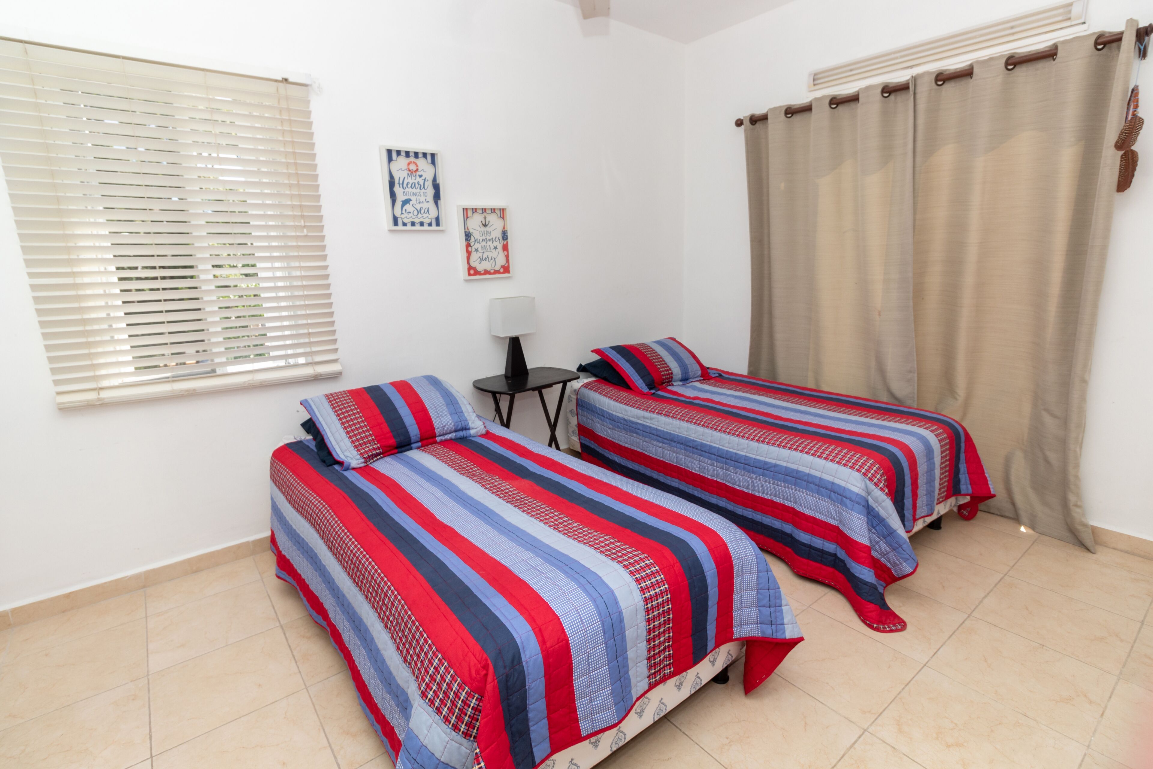 A bedroom with two beds, a window with blinds, a small table with a lamp, and a curtain.